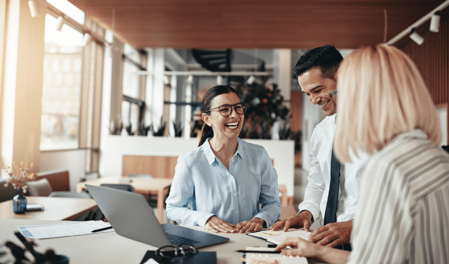 Laughing group of businesspeople working together on a laptop and going over paperwork while standing at a table in a modern office