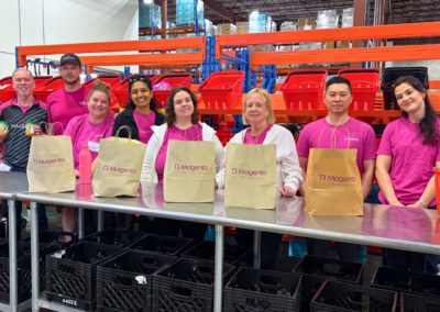 Group of eight volunteers in pink shirts standing behind a packing table with brown Magenta bags in a warehouse with red shelving bins in the background, smiling at the camera.