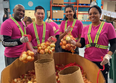 Four volunteers in pink shirts and safety vests sort and bag onions in a warehouse.