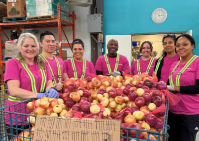 Group of volunteers in pink shirts and reflective vests pose around a large bin of onions in a warehouse.